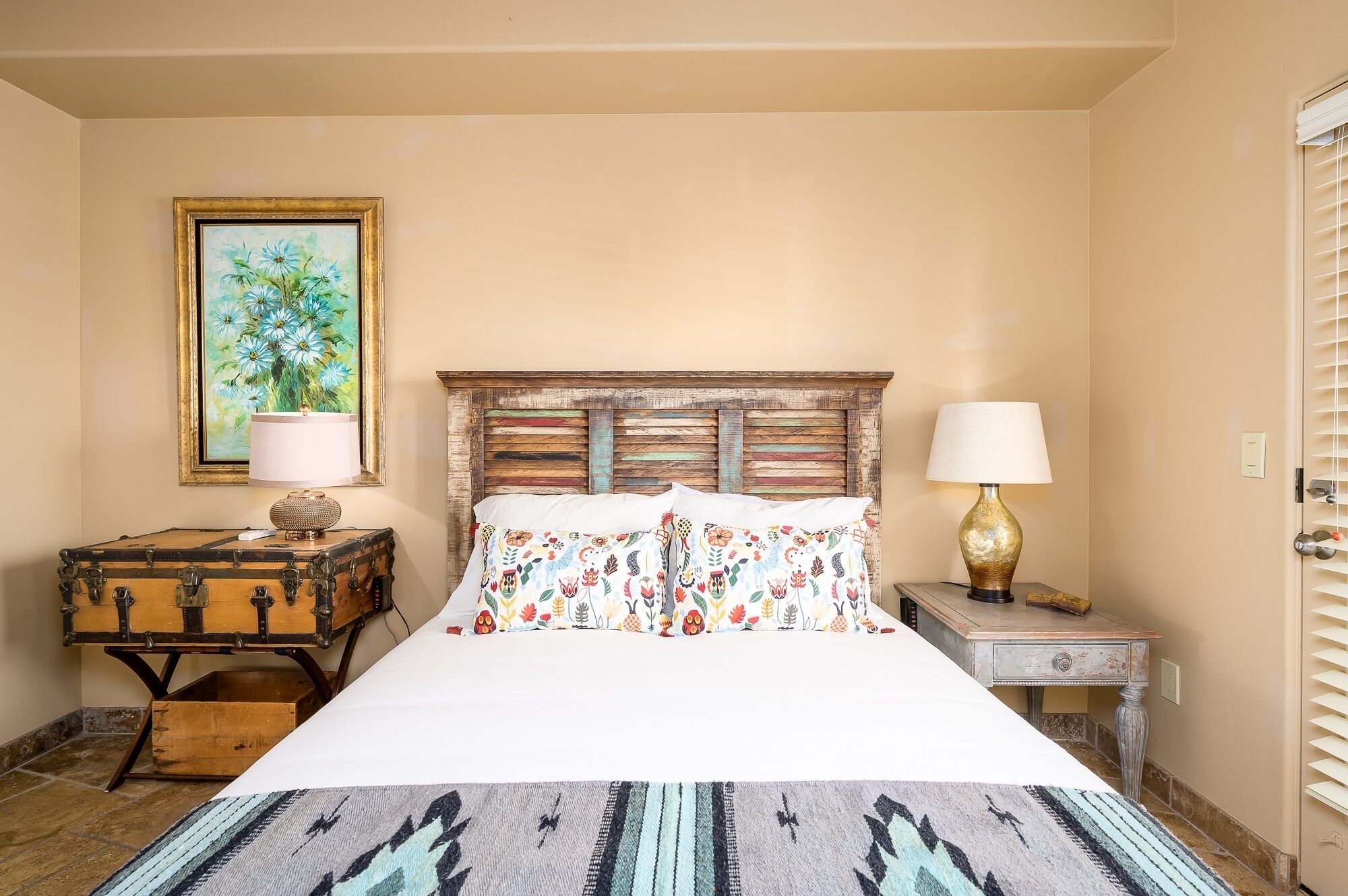 A bedroom with a wooden headboard, a bed with floral-patterned pillows, a bedside table with a lamp, a suitcase, and a framed painting on the wall.