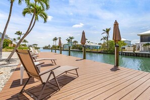 A wooden deck with lounge chairs and umbrellas, overlooking a calm body of water with palm trees and buildings in the background. - 21591/3 Waterfront Duplex With Pool And Spa - Manor 4 Bedroom Home (Fort Myers Beach)