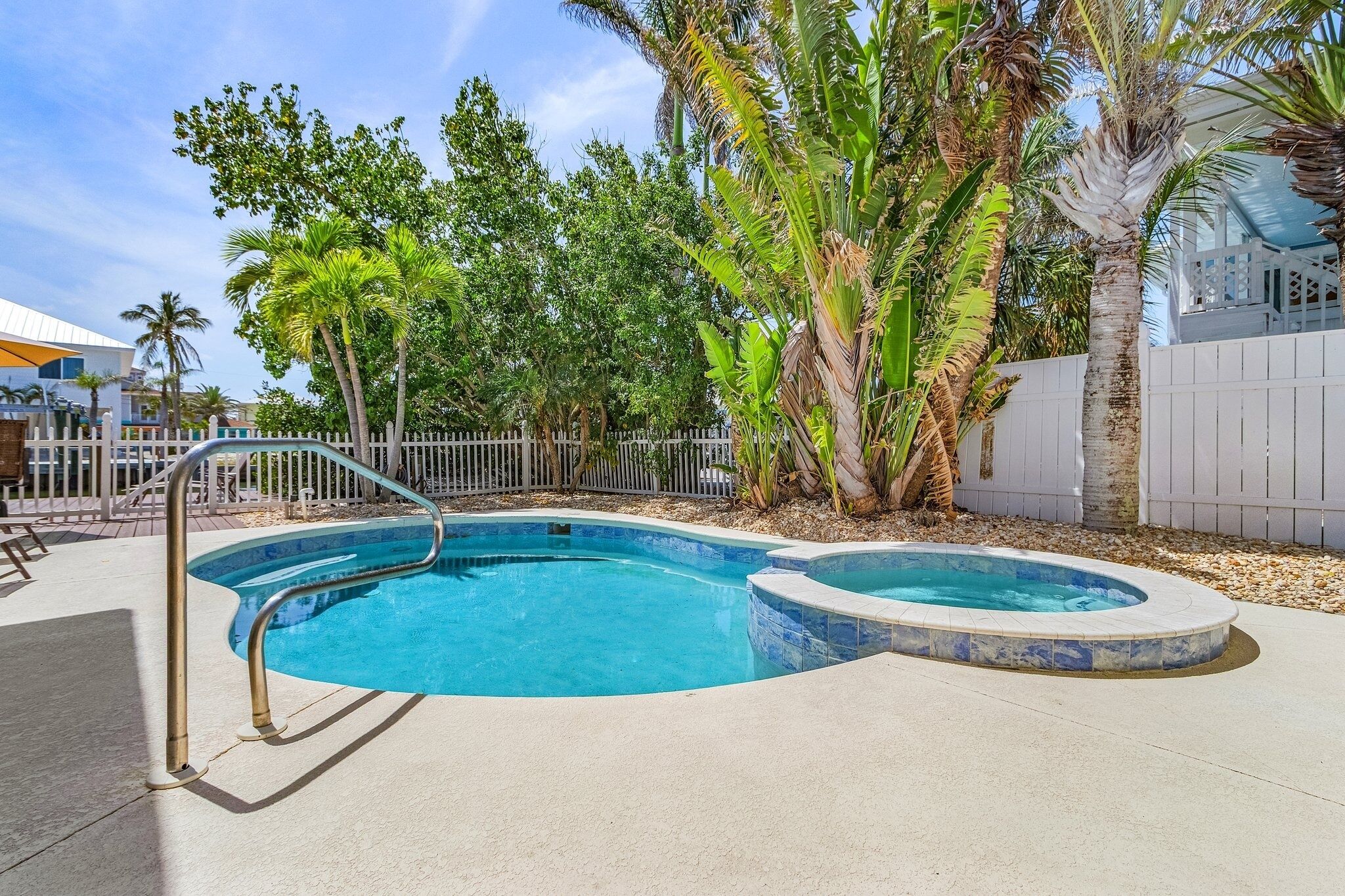 A swimming pool with a hot tub, surrounded by palm trees and a white fence.