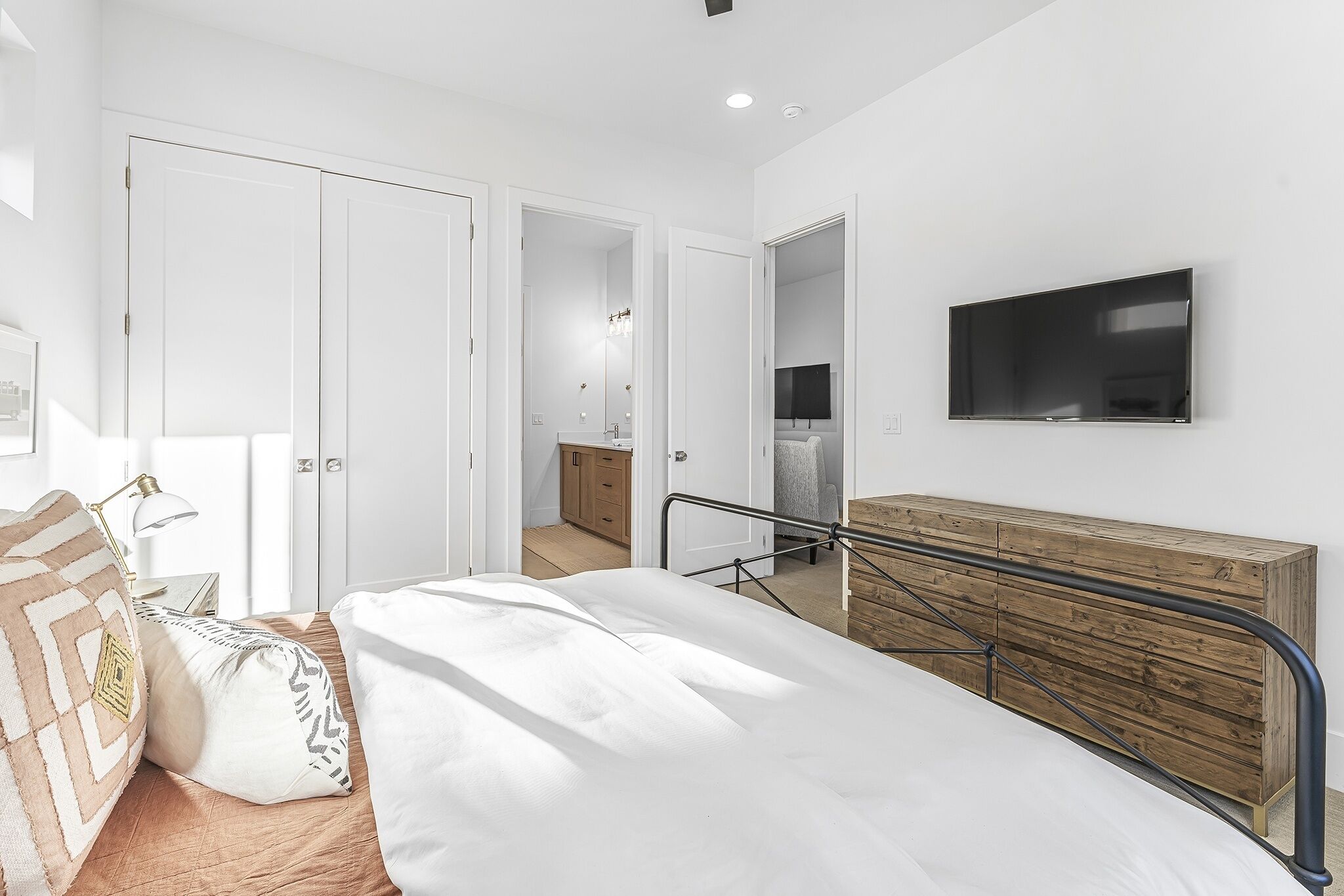 Une chambre à coucher moderne avec un lit, une commode en bois, un téléviseur fixé au mur et une salle de bain visible à travers une porte ouverte.