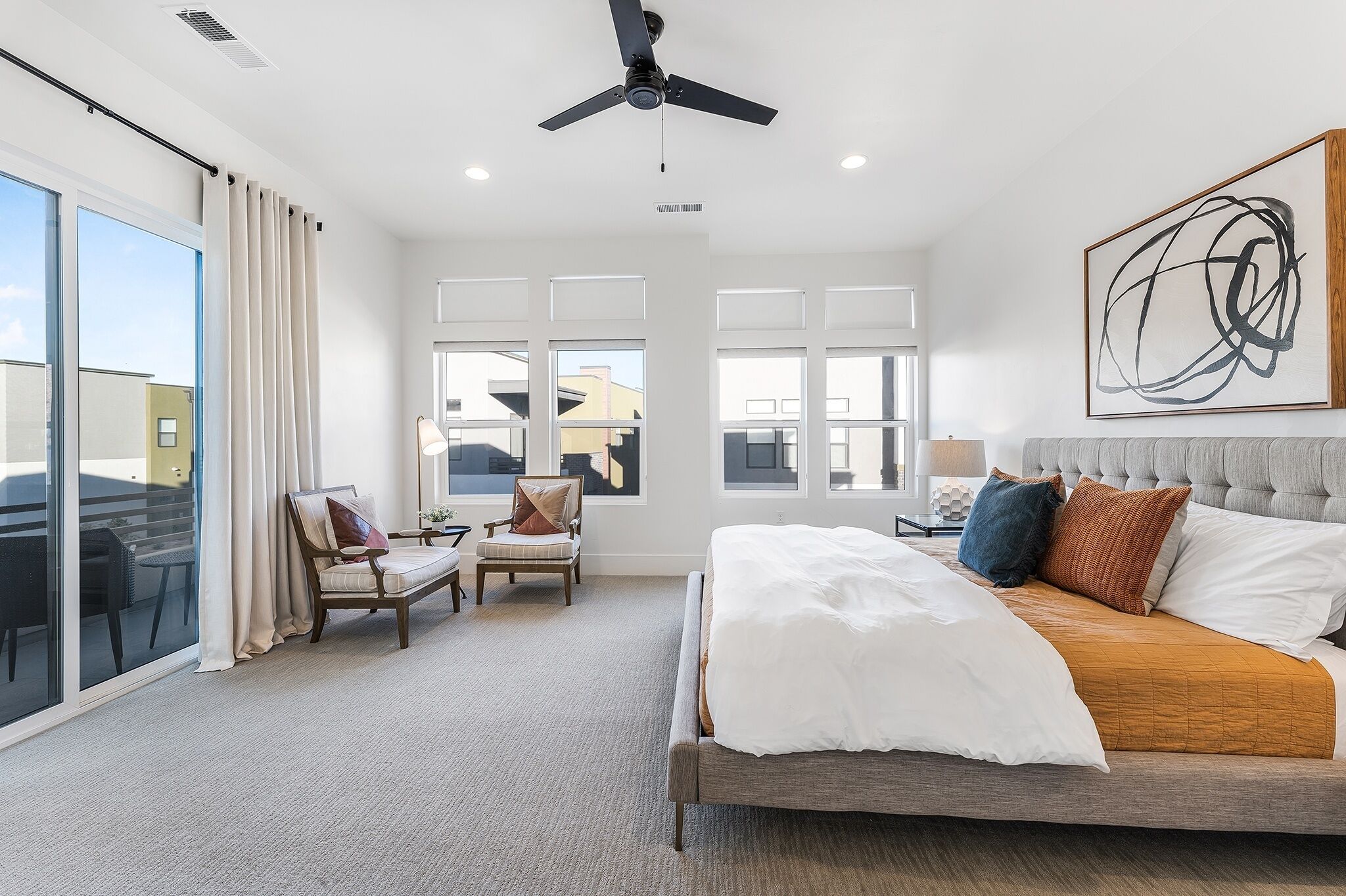 A modern bedroom with a large bed, a ceiling fan, a view of the city through large windows, and a seating area with a chair and table.