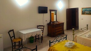 Un dormitorio con cama, una silla, una mesa con mantel, cómoda, espejo, televisor y puerta.