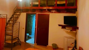 Habitación con escalera de madera, puerta que da a un baño, televisor sobre una repisa y un cubo de basura pequeño.