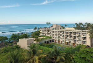 Un hotel frente a la playa con varios balcones, piscina y abundante vegetación.