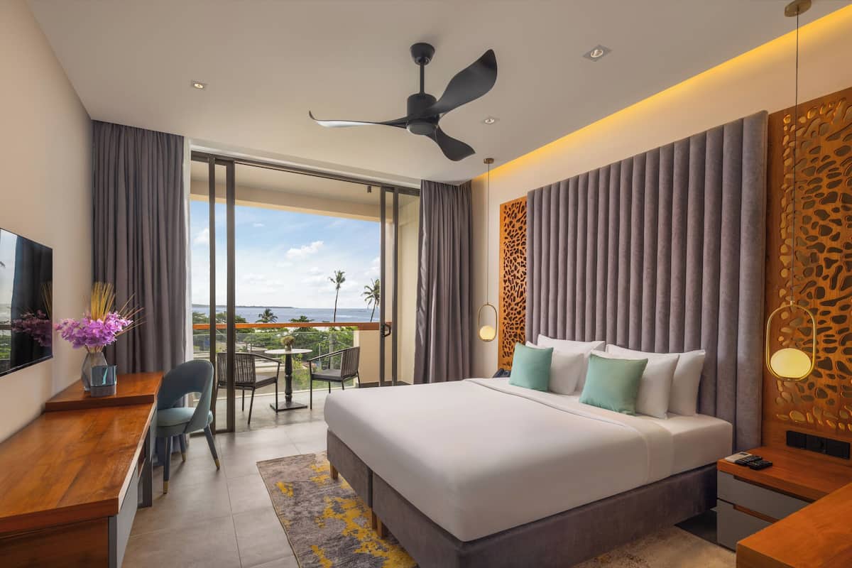 A modern hotel room with a large bed, a desk, and a balcony with a view of the ocean.