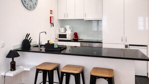 Et moderne køkken med hvide skabe, en sort bordplade, en stålvask, en mikrobølgeovn og en rød elkedel. Der er tre træbjerge og en sort lampe.