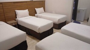 Habitación de hotel con cuatro camas individuales dispuestas en fila, cabecera de madera, mesita de noche con lámpara y refrigerador pequeño.