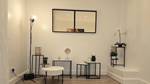 Una habitación con un suelo de alfombra a cuadrícula blanco y negro, una silla blanca, una pequeña mesa redonda con un espejo, una lámpara de pie y una orquídea en maceta.