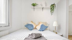 Una cama cuidadosamente hecha con un edredón a rayas, una toalla gris y dos almohadas en una habitación moderna con una pared blanca, una estantería con plantas y una lámpara de noche.