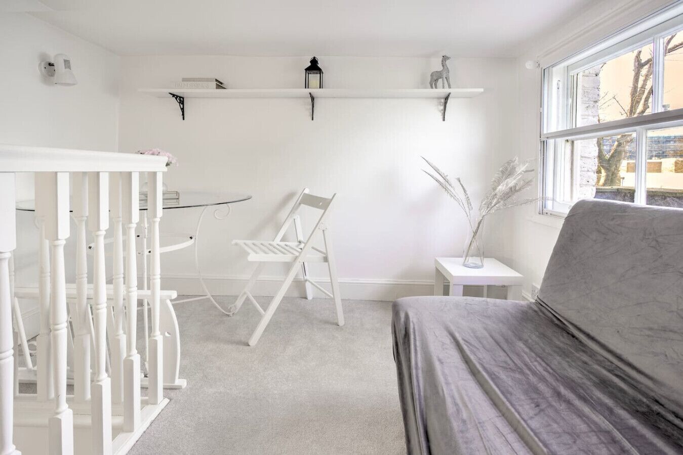 Une pièce minimaliste avec un berceau blanc, un canapé gris, une petite table et une fenêtre donnant sur des arbres.