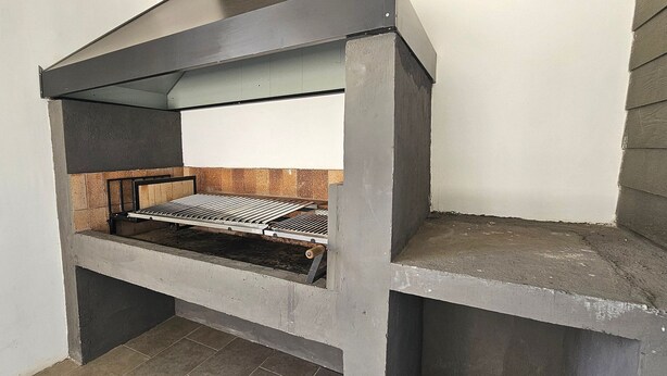 Ein kleiner, abgegrenzter Grillbereich mit einem Metallrost, einer Betonbasis und einer geneigten Dachkonstruktion.