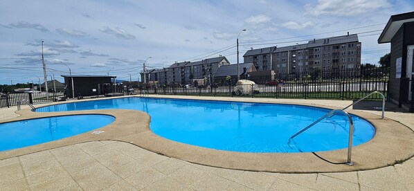 Piscina com design curvo, cercada por uma grade e um complexo de edifícios ao fundo.
