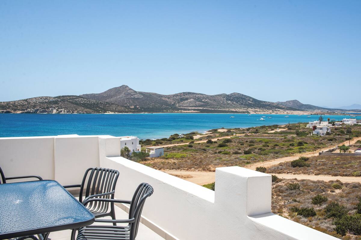 Une terrasse avec une table et des chaises donnant sur un paysage côtier, avec des montagnes et une mer bleue et limpide.