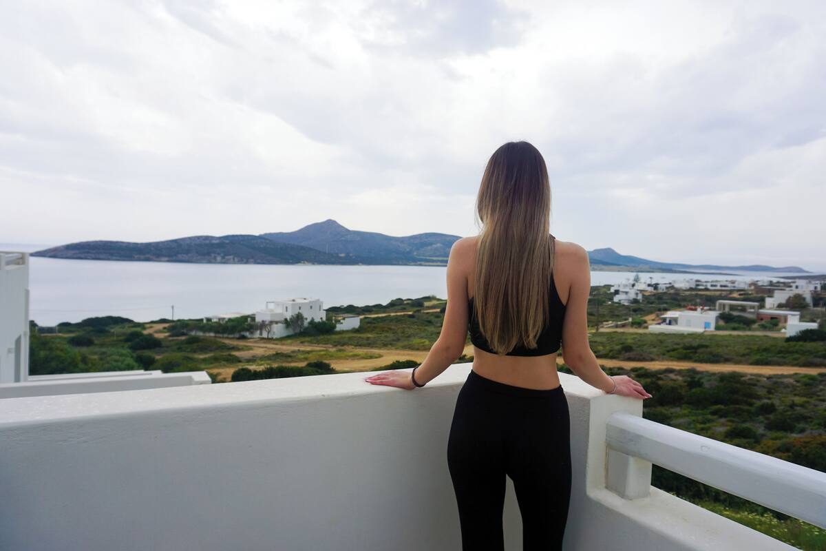 Une femme se tient sur un balcon d’où l’on peut admirer un paysage côtier, avec des bâtiments et des collines.