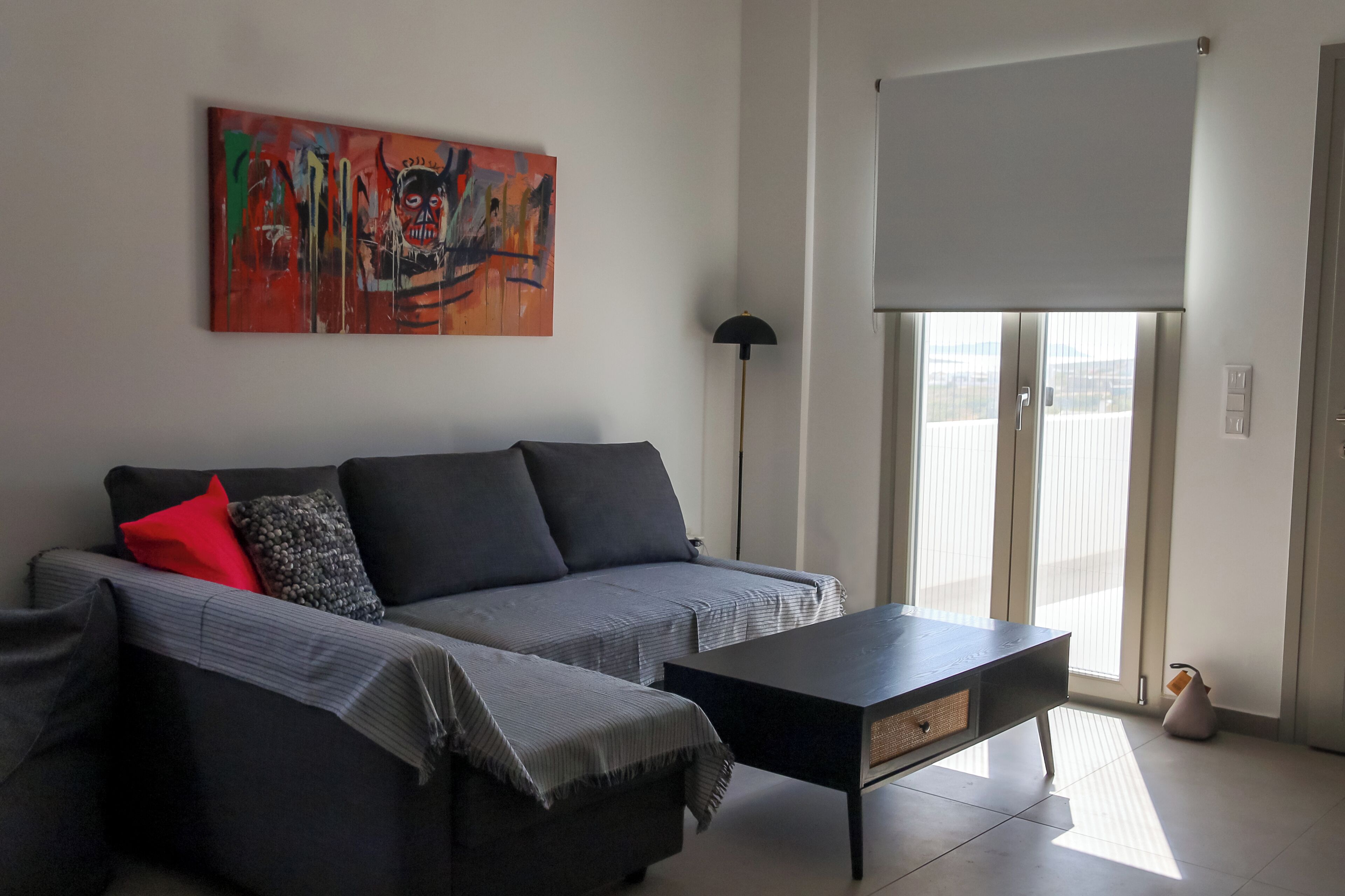 A modern living room with a grey sofa, a black coffee table, and a red pillow. There is a large abstract painting on the wall and a floor lamp.
