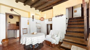 A room with a wooden staircase, a table with a striped tablecloth, a white armchair, and a rustic tiled floor.