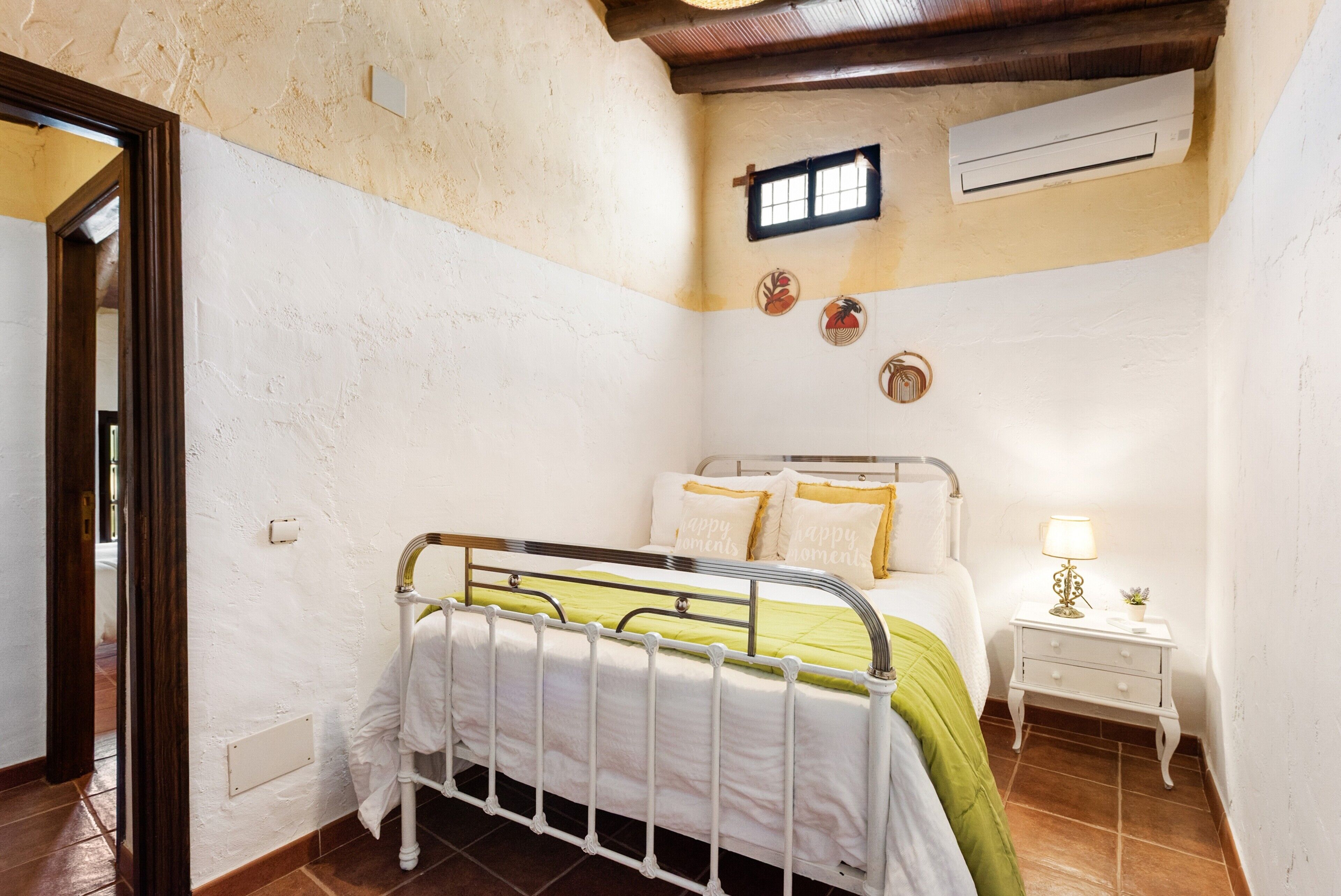 A bedroom with a metal bed frame, a bedside table with a lamp, and a small window.