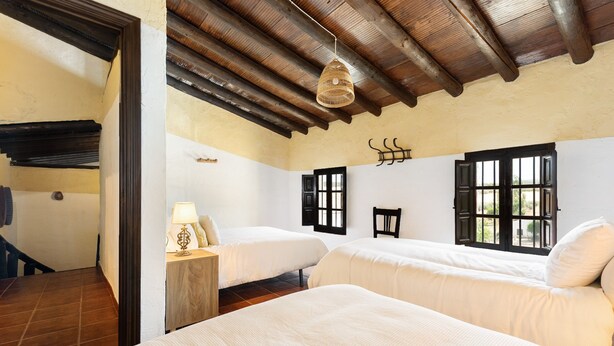 A room with two beds, wooden beams on the ceiling, a hanging light fixture, and a window with shutters.