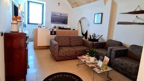 A cozy living room with a patterned sofa, a coffee table with magazines, and a rug with text. There's a kitchen area with a window and a cross on the wall. - baja delle sirene (Taranto)