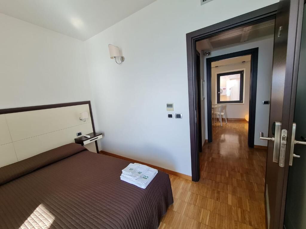 A hotel room with a bed, a bedside table, a lamp, and a door leading to another room.