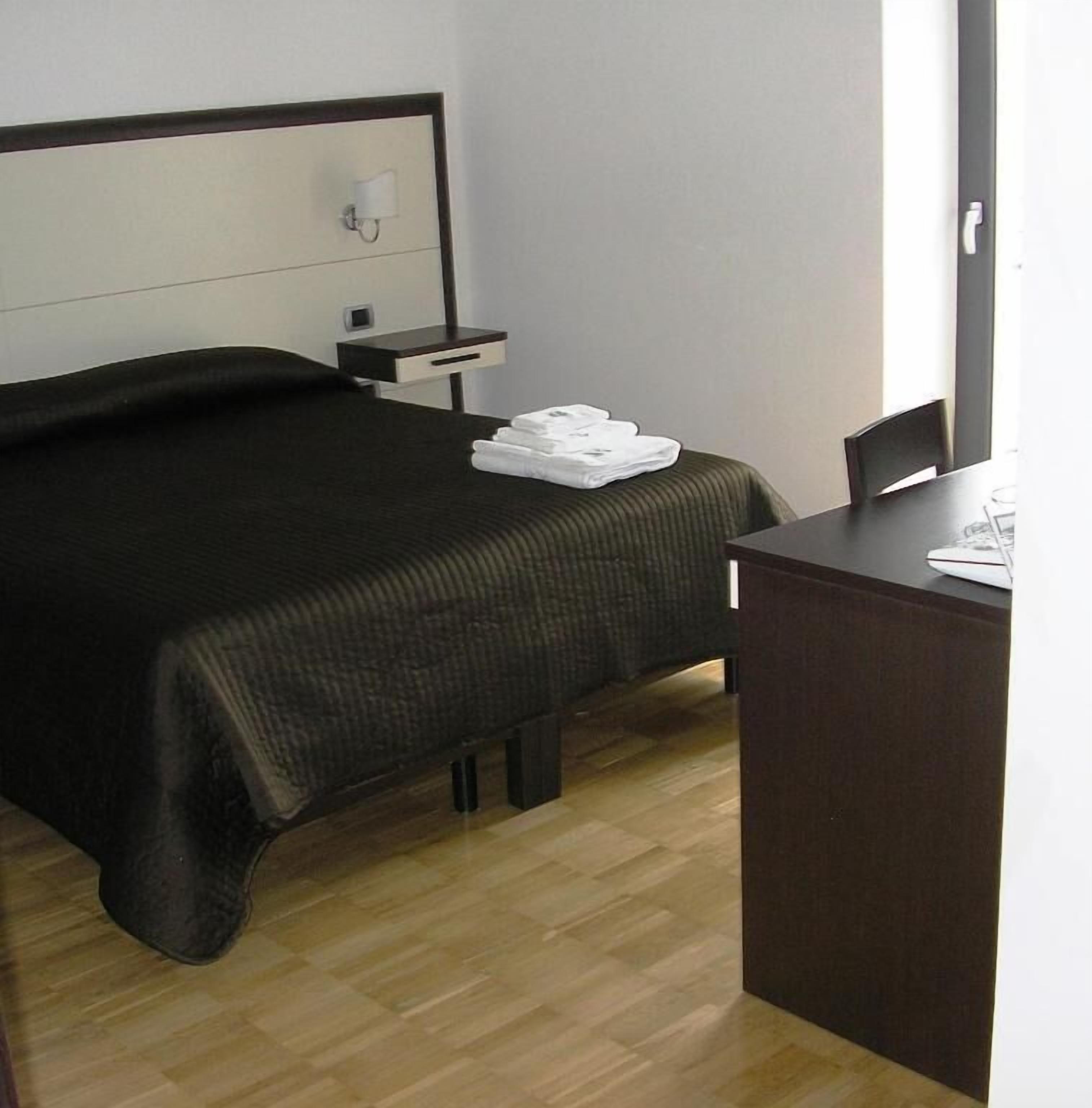 A hotel room with a bed, a bedside table with a lamp, a chair, and a desk with a chair.