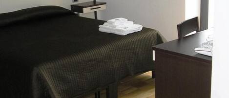 A hotel room with a bed, a bedside table with a lamp, a chair, and a desk with a chair.