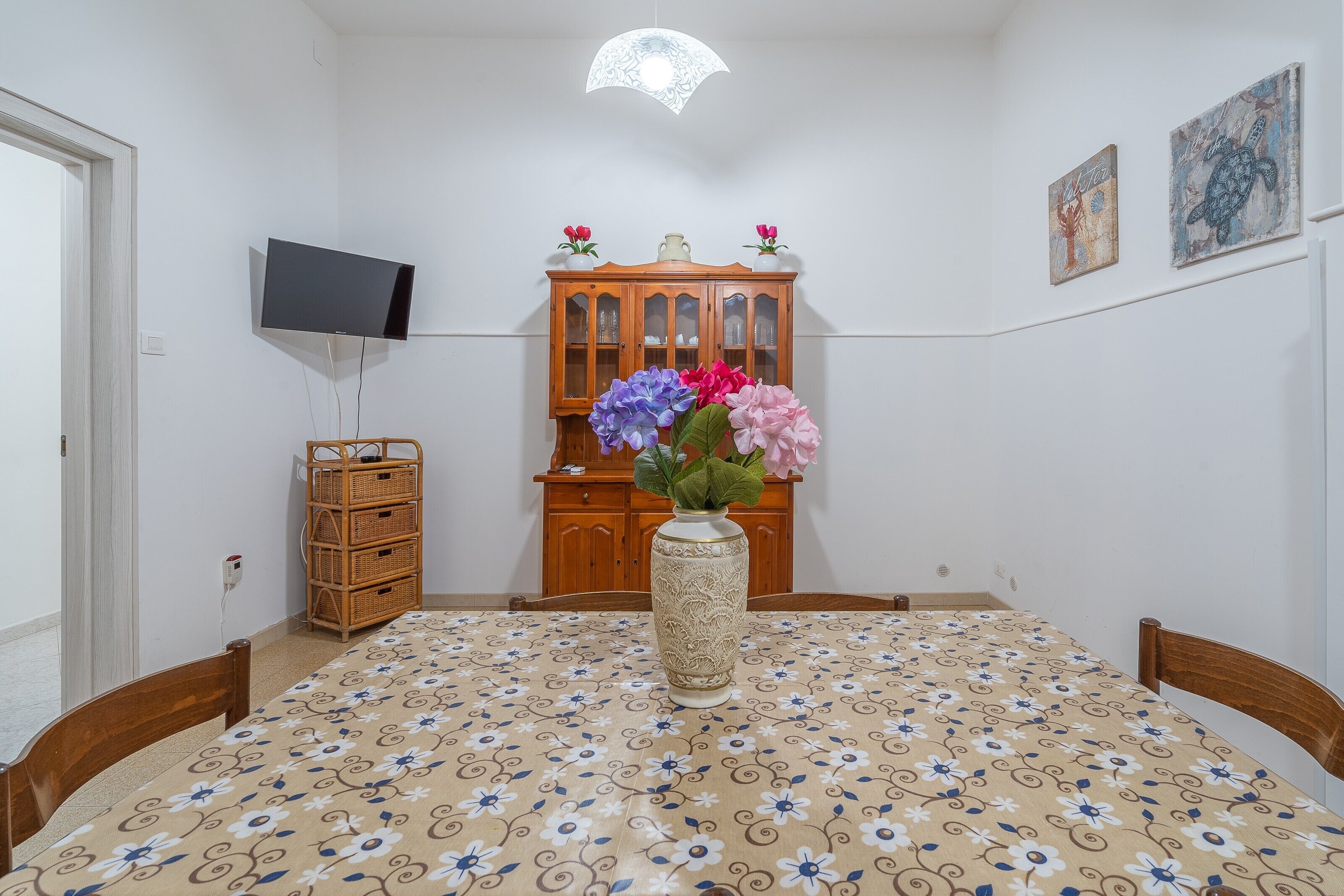 Une pièce comprenant une table ronde, un vase avec des fleurs, un téléviseur, un meuble en bois et des œuvres d’art murales.