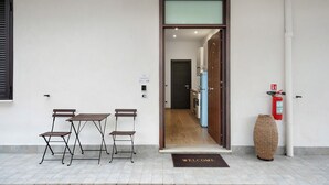 Entrada de un hotel con felpudo, extintor y un pequeño espacio de descanso con mesa y sillas.