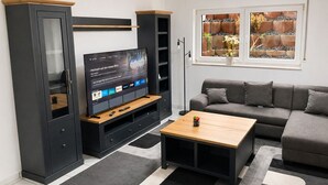 Un salon moderne doté d’un téléviseur à écran plat, de meubles aux couleurs sombres, d’un canapé gris et d’une fenêtre donnant sur un jardin.