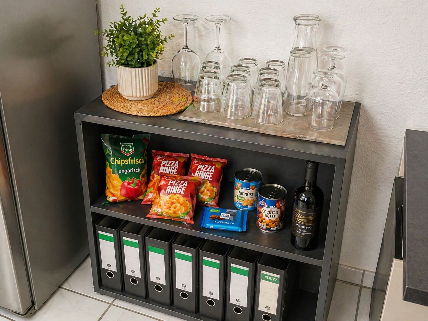 A shelf with snacks, a potted plant, wine glasses, and binders.