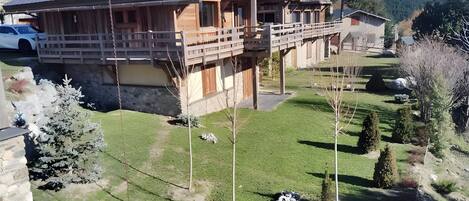 Una cabaña de madera con balcón, rodeada por un césped cuidado y un muro de piedra.