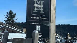 A sign for 'Le Hameau des Perles' with a wooden fence and a white van in the foreground.