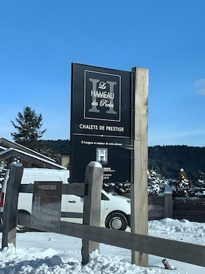 A sign for 'Le Hameau des Perles' with a wooden fence and a white van in the foreground.