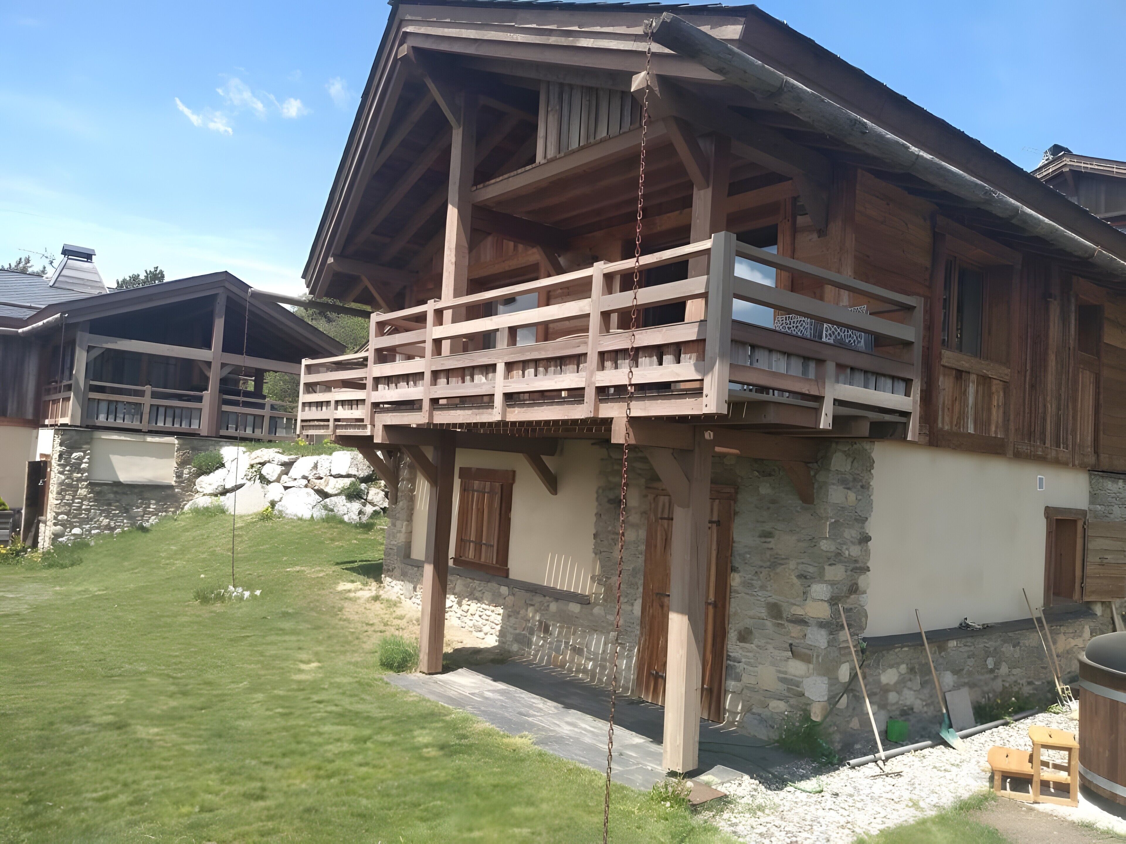 A wooden chalet with a balcony, stone walls, and a grassy yard.