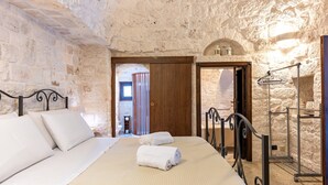 Ein Schlafzimmer mit Steinwand, einem Bett mit weißer Bettwäsche, einer Holztür und einem Metallregal.