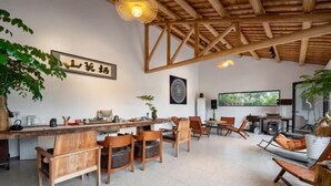 Una habitación amplia con techo alto y vigas de madera a la vista. Hay una mesa de madera larga con sillas, un sofá y una mesita con una planta.