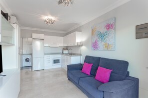 A modern kitchen with white cabinets, a built-in washing machine, and a grey sofa with pink cushions.