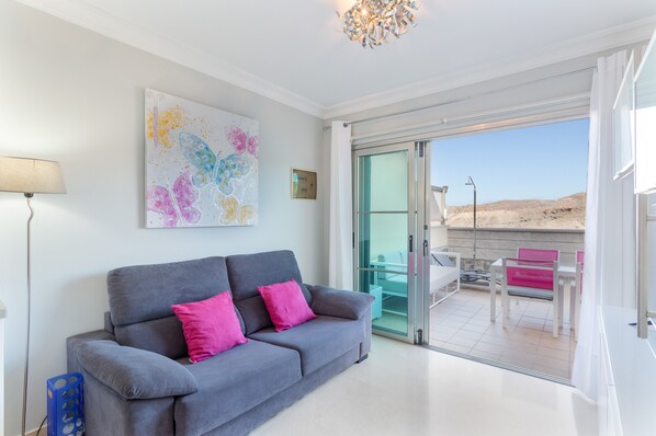 A modern living room with a grey sofa, a colorful abstract painting, and a glass door leading to an outdoor seating area.