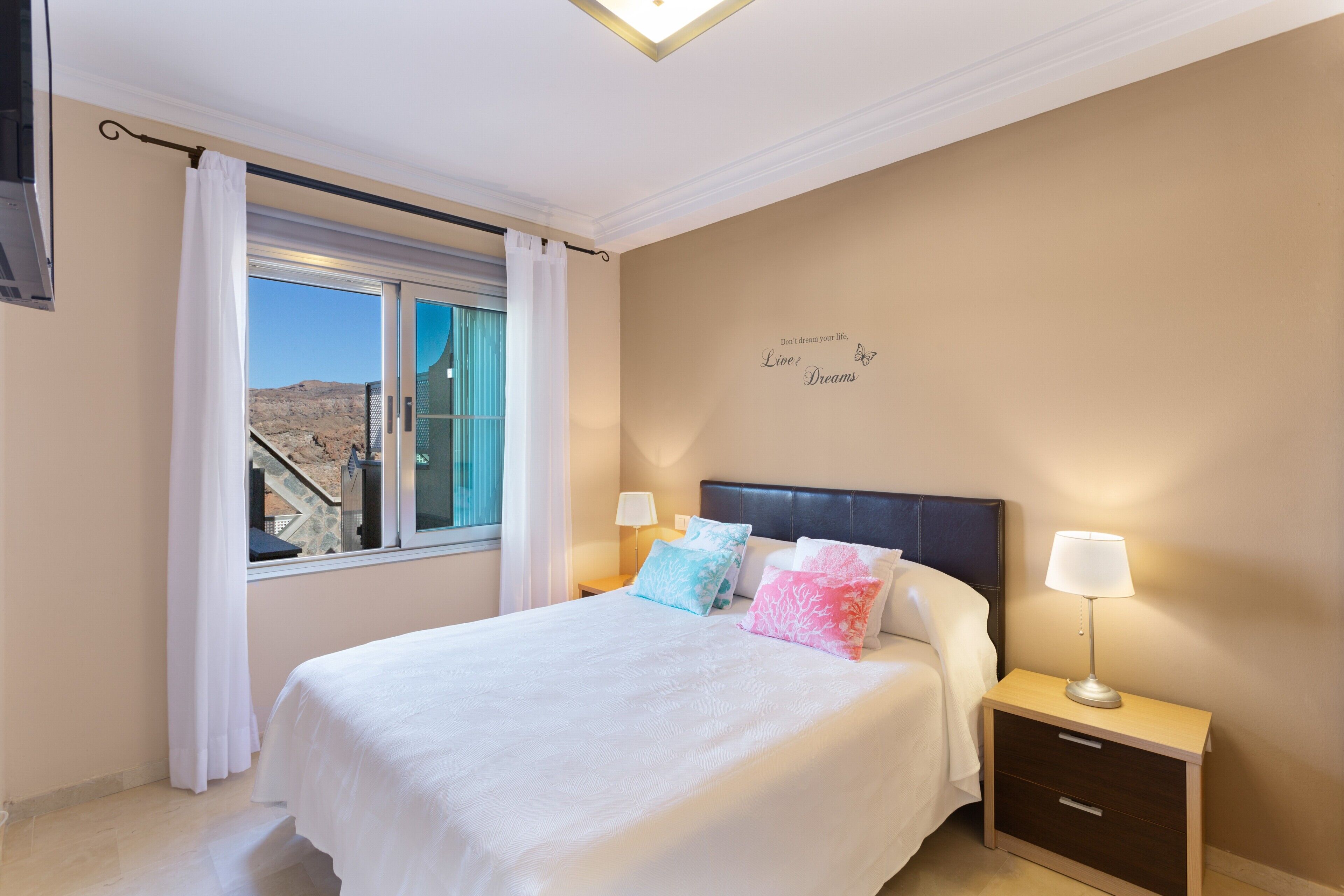 A bedroom with a bed, two bedside tables with lamps, a window with curtains, and a wall with a motivational quote.