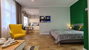 A modern hotel room with a bed, a yellow armchair, a dining area, and a TV on the wall.