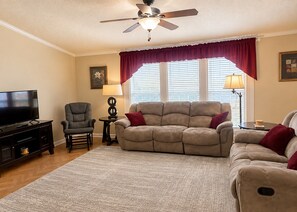 A living room with a flat-screen TV, a recliner, a sofa, a ceiling fan, and a window with blinds.