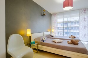 Habitación de hotel con dos camas, una mesita de noche con lámpara, una silla y vistas a un edificio a través de amplias ventanas.