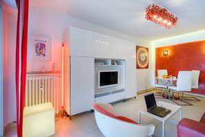 Una sala de estar moderna con un sofá blanco, una silla roja, una mesa de comedor con sillas, un televisor y una computadora portátil sobre una mesita.