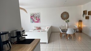 Et moderne køkken med kaffemaskine, et spiseområde med et hvidt bord og stole, og en opholdsstue med en hvid sofa og et dekorativt vægkunstværk.