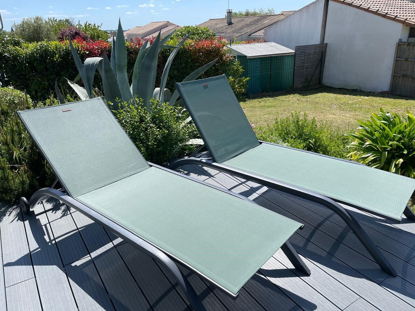 Zwei grüne Liegestühle auf einer Holzterrasse, im Hintergrund ein Garten und ein weißes Gebäude.