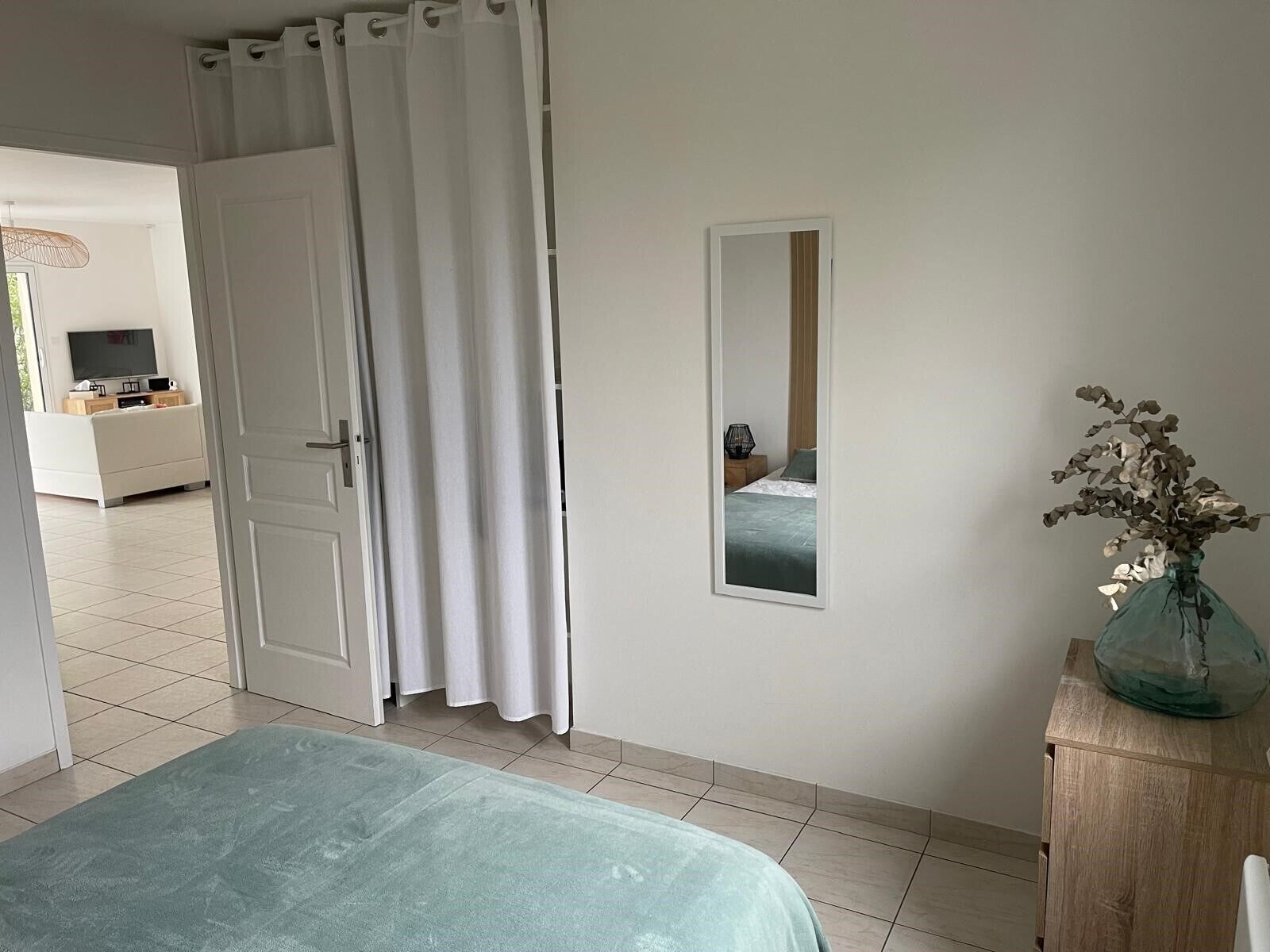 Una camera da letto con un letto, un comodino con un vaso, uno specchio e una porta che dà su un'altra stanza.