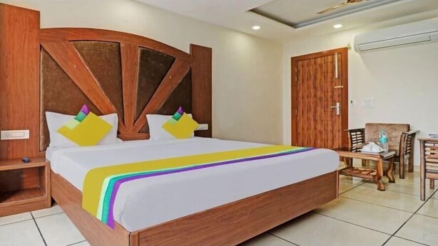 A hotel room with a large bed, wooden headboard, two bedside tables, a small table with chairs, and an air conditioning unit on the wall. - Hotel Grand Signia 9115357741 (Dera Bassi)