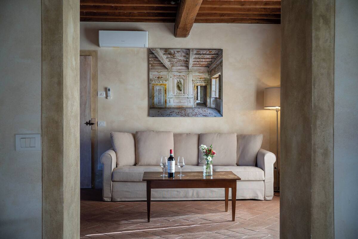 A room with a beige sofa, a wooden table with a wine bottle and glasses, a lamp, and a ceiling mural.