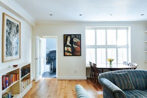 A spacious living room with a wooden floor, a blue sofa, a dining table with chairs, and a bookshelf filled with books. - Elegant Flat - Notting Hill. Minutes from Hyde Park. (London)