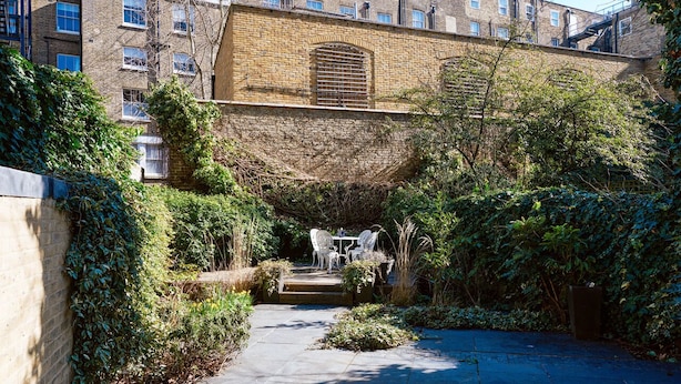 Un cortile con un muro in mattoni, un piccolo tavolo con sedie e fitta vegetazione.
